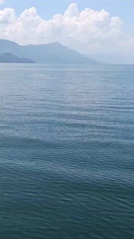 A boat sails across a large body of water. In the distance, hazy mountains rise above the horizon. The sky is partly cloudy. The water is blue and appears calm with gentle ripples. The boat is moving from left to right across the frame.