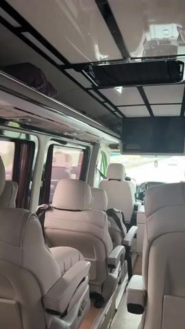 The camera is inside a van with plush white leather seats, viewed from the back, facing toward the front. A driver, seen from behind, is operating the van on a highway. A TV is mounted to the ceiling. There are overhead storage compartments. The interior of the van appears luxurious. The light is bright.