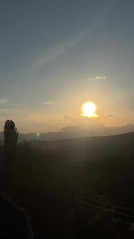 The video shows a sunset over a mountainous landscape. The sun is bright and partially obscured by clouds. Below the mountains is a village or town with houses visible among the trees. Power lines run across the lower part of the frame in the foreground. As the video progresses, the sun sets further, the sky lightens, and more of the houses and landscape become visible.
