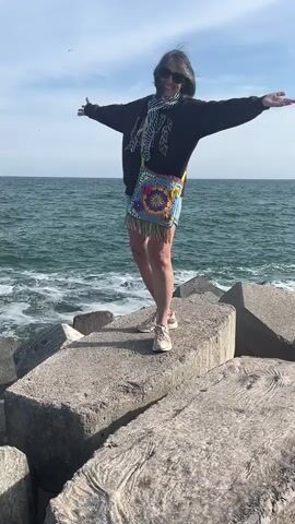 A woman with gray hair and sunglasses stands with her arms outstretched on large concrete breakwater blocks at the seaside in Palavas-les-Flots, France. She is wearing a dark long-sleeved top, a striped scarf, denim shorts, and light-colored sneakers. A vibrant, colorful crocheted bag with a floral pattern and fringe hangs across her body. The woman is smiling, appearing joyful as she poses against the backdrop of the ocean. The sea is choppy with visible waves, and the sky is bright and partly cloudy, indicating a sunny day. The overall scene depicts a happy moment on a coastal breakwater.