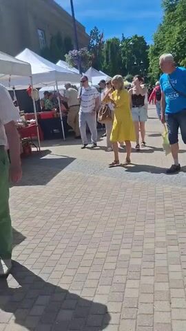 The video captures a sunny outdoor market scene. In the foreground, a man in a blue t-shirt and shorts walks towards the right, carrying a light-colored tote bag. To his left, a woman in a yellow dress walks in the same direction. Behind them, a woman in a white top and beige pants walks towards the left, and a young woman in a black lace top and denim shorts also walks towards the left. Several market stalls with white canopies are visible in the background, along with trees and buildings. Other people are browsing the stalls and walking through the market. The ground is paved with brick-like stones.