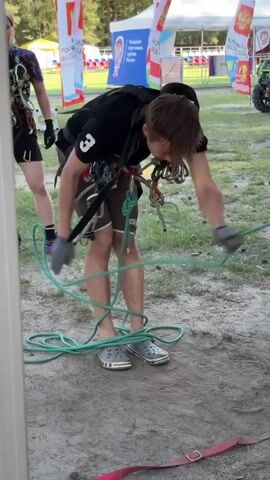 The video shows a young man and a young woman outdoors, preparing for an activity involving climbing ropes and harnesses. The man is wearing a black t-shirt with the number "3" on it, brown shorts, and grey Crocs. He has a complex harness with multiple ropes and carabiners attached. He is bent over, actively managing a coiled green rope on the ground, seemingly preparing it for use. The woman, standing to his left, is also wearing a harness with ropes and carabiners, suggesting they are both participating in or assisting with an outdoor adventure or training.

The setting appears to be a park or an open field with grass and sandy ground. In the background, several large white flags with red and blue elements are visible, along with some tents and structures that suggest an organized event or competition. One of the flags has text in Russian, which appears to be related to the "All-Russian Sports Competition". The flags and background suggest the event is taking place in Russia, specifically in the Penza region, given the context.

The time of day appears to be daytime, likely afternoon, given the natural lighting and shadows. The weather seems pleasant for outdoor activities. The individuals' focused expressions indicate concentration on the task at hand. The overall scene conveys a sense of preparation for an athletic or adventurous activity, possibly related to climbing, rappelling, or a similar rope-based sport.
