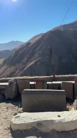 The video shows a collection of broken, carved stone blocks set against a mountainous backdrop under a bright, clear blue sky. The stone blocks are varied in shape and size, some clearly fragments of larger structures, others seemingly intact but lying scattered.  A weathered wooden pole stands among the stones, bisected by a thin white wire running diagonally across the upper portion of the frame.  The mountain in the background is predominantly brown and sparsely vegetated, suggesting a dry, arid climate. The overall scene suggests an archaeological site or a collection of ancient ruins.