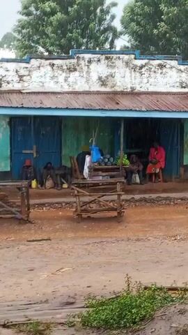 The video depicts a street scene in a village during heavy rainfall. A line of people are sitting under the porch of a building with a dilapidated facade featuring white, blue, and turquoise paint. The ground is muddy and there are wooden structures present. In the background, trees, utility poles, and neighboring buildings can be seen through the rain. The atmosphere appears bleak and wet.
