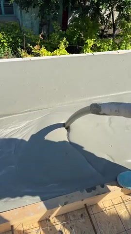 A concrete pump hose is pouring liquid concrete into a formwork. The concrete is a light gray color and appears to be flowing smoothly. The formwork is made of wood and is supported by rebar. In the background, there are green trees and a building. The sun is shining, casting shadows on the concrete and the surrounding area. There is a shadow of a person holding the concrete pump hose.