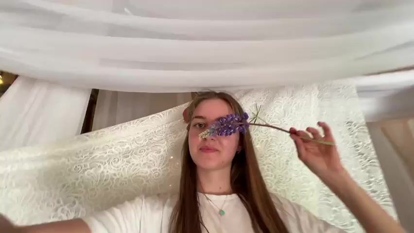 A young woman with long brown hair holds a purple lupine flower in front of her eye. She is positioned in front of draped white fabric, some of which is lace. Behind the fabric, wooden beams and a wall can be seen. The woman is looking at the camera and smiling slightly. She quickly moves the flower away from her face.