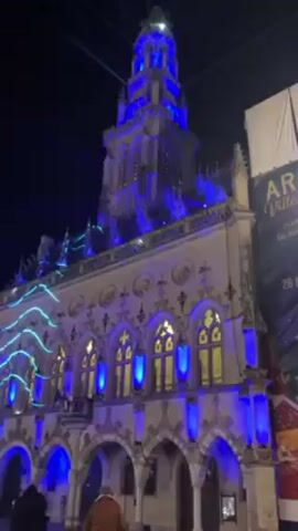 At night, an ornate stone building is illuminated with blue lights outlining its structure and windows. A string of wavy, pale blue lights runs across the top of the building. A tall tower, also lit in blue, rises from the top. Two people walk in front of the building; one wears a brown jacket. A poster is visible on the right side of the frame.