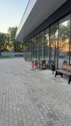 A young child, wearing a pink shirt and blue shorts, walks along a paved walkway in front of a modern building with large glass windows. The building has a curved, gray overhang. Trees and a clear sky are visible in the background. The setting appears to be outdoors, possibly near a commercial establishment, during the golden hour as the light suggests sunset.