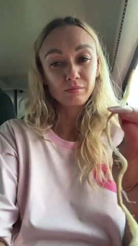 A woman with blonde hair is holding a long, curly, yellowish-white food item. She puts it over her face, using it as a fake mustache. She is wearing a light pink shirt with a heart on it and the word "Milk". She is sitting in a car. The inside of the car and the window can be seen in the background.