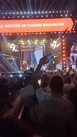 The video captures a live concert from the perspective of an audience member. The foreground is filled with the backs of numerous people, many of whom are holding up their phones to record the event. Above the crowd, the stage is brightly lit, displaying the singer Derkan Kaya on a central screen, flanked by his name and the logo of the "Kültür Yolu Festivali." Kaya is also visible in person performing live on stage with his band. The stage is framed by a large, rectangular structure, adorned with rows of bright lights. The overall atmosphere is energetic, suggesting a well-attended and enthusiastic concert.