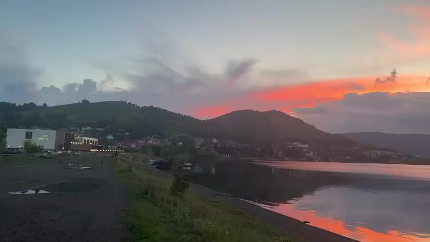This video captures a serene sunset over Lake Yakty-Kul in Russia. The scene is dominated by a tranquil lake reflecting the vibrant hues of the twilight sky. Hills covered in lush greenery rise from the far shore, dotted with small houses and buildings that form a village nestled along the water's edge. The sky is a canvas of soft blues, pinks, and oranges, with dramatic clouds adding texture and depth to the sunset.

In the foreground, a gravelly shore with some patches of grass leads down to the water. A few small boats are moored near the shore, suggesting activity on the lake. A modern building with a sign featuring a green logo is visible on the left side of the frame, possibly a hotel or resort. The overall atmosphere is peaceful and picturesque, evoking a sense of calm and natural beauty. The time of day is clearly evening, as indicated by the sunset. The weather appears to be calm, with a gentle breeze creating subtle ripples on the water. The location is identifiable as Yakty-Kul, Russia, through its distinct landscape of hills, a lake, and a settlement. No discernible text other than the logo on the building is visible.