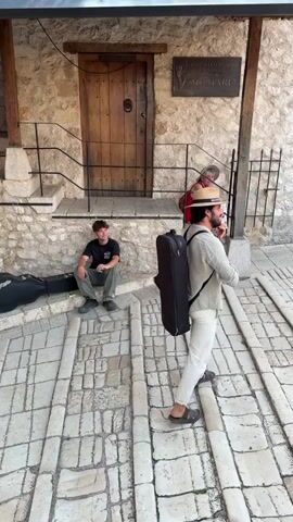 The video opens on a cobblestone path leading up to a stone building with a wooden door and a sign above it. A person wearing a straw hat and a light-colored outfit, carrying a long black case, walks away from the camera while playing a wind instrument. To the left, another person sits on the steps of the building. The camera then pans to the right, showing a woman in a white outfit walking down the cobblestone path, followed by a man in a t-shirt and shorts carrying a backpack and holding a snake. In the background, there are more stone buildings, a minaret, and several people on a balcony. The final shots show a blurry view of people on a balcony.