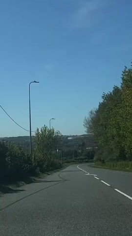 The video shows a front-facing view from a vehicle driving down a winding road on a bright, sunny day. The road, which appears to be asphalt, curves gently to the right and descends into a valley. On the left side of the road, there are tall lampposts and dense green trees and bushes, with some bare branches indicating early spring or late autumn. A red triangular road sign is partially visible on the left, close to the edge of the road, though its specific text is unreadable. On the right, the road is bordered by a line of trees with green foliage.

In the distance, a sprawling settlement or town can be seen under a clear blue sky, extending across the horizon. The sun casts distinct shadows on the road surface, suggesting midday.

A prominent detail is the word "SLOW" painted in large white letters directly onto the road surface, indicating a need for reduced speed. White road markings, including a solid line and a dashed line, are visible on the right edge of the carriageway. The overall scene suggests a drive through a semi-rural area transitioning towards a populated zone in South Kesteven, United Kingdom. The sound of an engine hums throughout the clip.