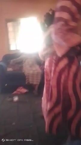 The video shows the interior of a room with orange walls and a bright window. A person in a striped dress is draped over a dark blue couch. A person in a light-colored outfit is seen standing by the wall, then walking towards the couch. The floor has a black rug with items scattered on it, including sandals, a pink garment, and a green bowl. The room appears somewhat cluttered.