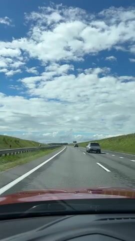 A first-person perspective video captures a view from the driver's seat of a vehicle traveling on a highway. The road ahead is paved and marked with white lines. Green grassy hills flank both sides of the highway, separated by guardrails. The sky is a vibrant blue, dotted with fluffy white clouds. Several vehicles are visible on the road, including a silver car in the right lane and two smaller vehicles further ahead. The driver's dashboard is partially visible in the foreground, with a portion of a red vehicle's hood. The scene suggests a daytime journey under clear, cloudy skies.