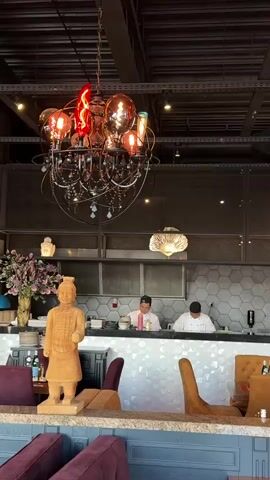 The video captures the interior of a restaurant. In the foreground, a low-angle shot reveals plush purple seating and a textured counter. A terracotta warrior statue stands prominently on the counter, accompanied by a vase of flowers and a lamp. In the mid-ground, a sleek bar area is visible, with two chefs in white uniforms working behind it. One chef is wearing a white hat, and the other a black baseball cap. They appear to be preparing food. Behind the bar, a wall with a hexagonal pattern is visible. Above the bar, an ornate chandelier with multiple light bulbs, some emitting a red glow, hangs from the ceiling. The ceiling itself is industrial, with exposed metal beams and ductwork.