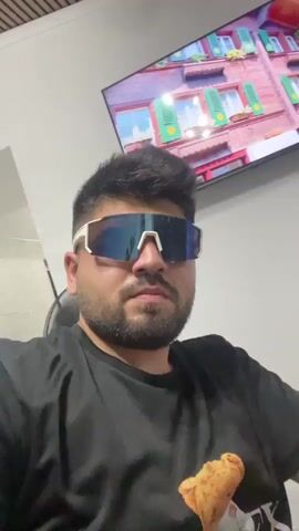 A medium shot captures a man with dark hair and a beard wearing white-rimmed sunglasses, a black t-shirt with a graphic of a teddy bear in an astronaut suit, and a white t-shirt partially visible underneath. He is seated, looking directly at the camera. Behind him, a television screen displays a colorful image of buildings with green shutters. The environment appears to be indoors, with a white wall and a glimpse of a black chair. The man's left arm is visible resting on his lap.