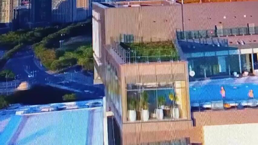 An aerial shot shows a modern hotel with a large "X" logo on its side. The hotel has glass windows and a green rooftop garden. In the background, a body of water with several sailboats is visible, along with trees and other buildings. The video pans slightly, offering a wider view of the hotel and its surroundings.