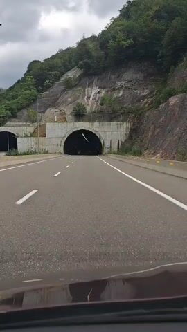 A car drives down a paved highway towards a large, arched tunnel. The tunnel is carved into a steep, rocky mountainside covered with green trees. The sky is overcast and cloudy. The highway is marked with white lane lines and yellow reflectors along the side. The tunnel entrance is reinforced with concrete structures. The interior of the tunnel is dark.