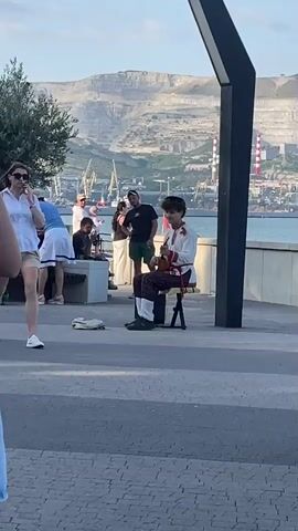 A street musician plays an instrument while sitting on a stool in an outdoor setting. He is wearing a white shirt with red and black trim and plaid pants. A woman walks by in the foreground, talking on a cell phone, wearing a white shirt and tan shorts. Other people are visible in the background, some standing and some sitting on a bench. There are trees, a body of water, cranes, industrial buildings, and a mountainous landscape in the distance. The ground is paved with interlocking stones. The lighting suggests it is daytime.
