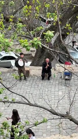 The video shows a pair of musicians performing outdoors on a cobblestone surface. One musician, wearing a black outfit, sits on a stool and plays a wind instrument. The other, dressed in a camouflage jacket and dark pants, stands to his left playing a frame drum. An amplifier sits on a small stand nearby. Several people are walking in the vicinity, some glancing at the musicians. The scene is framed by tree branches with fresh green leaves. Several cars are parked in the background.