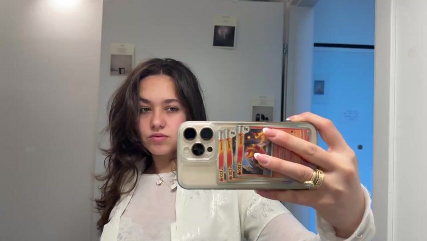 A woman takes a selfie in a bathroom mirror. She is wearing a white top with a white patterned jacket. She has long, wavy brown hair. Her phone case has Pokemon cards stuck to it. She runs her hand through her hair. The camera pans over to a man standing in a room. He is wearing a tan jacket with decorative straps, a plaid shirt, and a black t-shirt. The room is decorated with various pictures and posters on the walls. There is a television, a couch, and a desk visible in the background. The lighting in the room is blue.