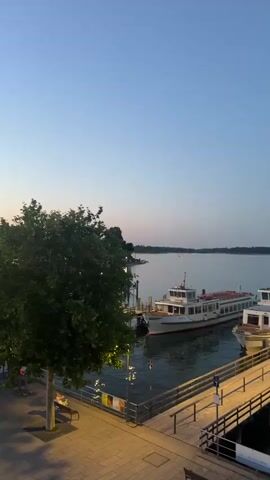 The video shows a twilight view of a lake with several passenger boats docked at a wooden pier. In the foreground, a paved walkway runs alongside a treed area, with benches visible. The sky is a deep blue, transitioning to a lighter hue near the horizon. The water is calm, reflecting the soft light and the lights from the pier and boats. One boat is white with red trim, and another is white with green trim. The pier has wooden planks and metal railings, with some light fixtures along its length. The overall atmosphere is serene and peaceful.