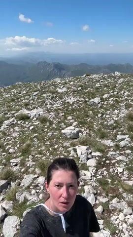 A woman with shoulder-length dark hair is recording a video. She is standing in a rocky, mountainous terrain with scattered patches of green grass. In the background, there are distant mountains and a clear, blue sky with some clouds. As she moves the camera, two other people are visible in the background, sitting on rocks. Later, they walk toward the camera. The woman smiles and waves at the camera. The camera's orientation is vertical.