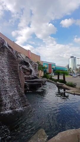 Here is a description of the video:

The scene depicts a man-made waterfall and pond, likely part of a landscape design near a commercial area. The waterfall is constructed against a brick wall, appearing like a series of rocky steps that lead water into the pond below. The pond's water is dark, possibly due to its depth or the surrounding shadows. In the background, there is a road with some traffic and commercial buildings. The sky is blue with scattered clouds, indicating a sunny day. Some greenery and palm trees are planted around the waterfall and pond. The camera appears to be stationary.