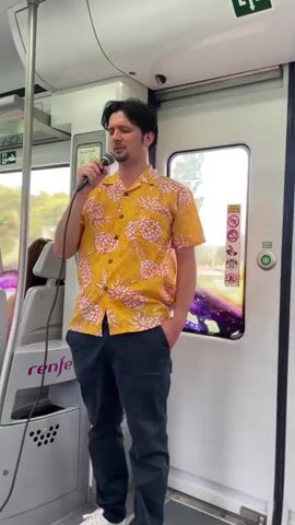 A man wearing a yellow pineapple-print short-sleeved shirt and dark pants holds a microphone in his right hand and sings. He is standing on a train, facing forward. The train interior is visible, with gray seats and poles. A window with an abstract, colorful image is to the right of the man. The word "renfe" is visible on the train's interior. The man's left hand is in his pocket.