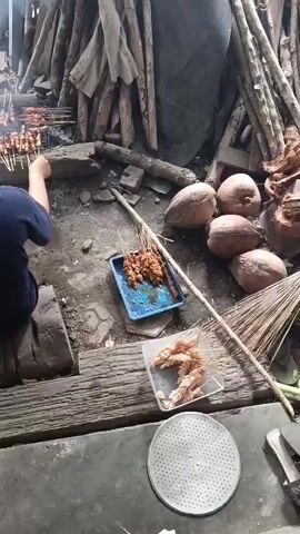 A person is sitting on the ground in front of a grill. They are wearing a dark blue shirt and dark blue shorts. They have their hair pulled back in a bun. The person is reaching towards the grill with their left hand. On the grill, there are several skewers of meat being cooked over a fire. There is smoke rising from the grill. In front of the person, on the ground, are two blue trays filled with skewers of meat. To the right of the person, there are several coconuts and a broom. There is also a white circular object with holes in it, possibly a sieve or a grill grate. The ground is dirt and there are wooden planks laid out. The background consists of a structure made of wooden logs and some sort of material covering them.