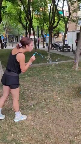 The video captures a playful daytime scene in a park in Sant Just Desvern, Spain. A woman with light brown hair, wearing a black tank top, black shorts, and white sneakers, stands on a patch of dry grass and dirt, actively blowing bubbles from a small wand.

A brown and white dog, resembling a Border Collie, runs into the frame from the right, immediately focusing on the bubbles. The dog excitedly jumps in an attempt to catch the floating bubbles, its tongue slightly out, indicating its playful engagement. It then lands and continues to look around, anticipating more bubbles.

The setting is a public green space with several trees providing shade. In the background, multi-story brick buildings with windows are visible, suggesting an urban or suburban environment. One building clearly displays signs for businesses, with "MOBLER" (likely "Muebles" for furniture) and "Estetica" readable. Other background elements include streetlights and benches where people are seated, reinforcing the public park setting. The weather appears sunny and clear, contributing to the lighthearted atmosphere of the interaction between the woman and the dog.