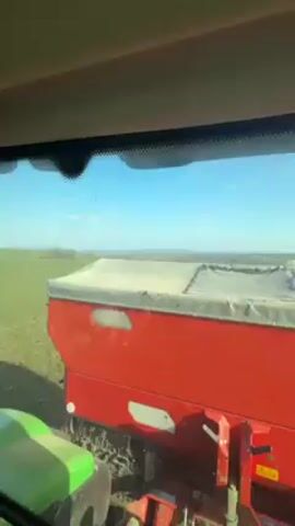 The video shows the view from inside the cab of a John Deere tractor while it is driving through a field. The tractor is pulling a red seeder implement behind it. The field is green with rows of planted crops. A mountain range can be seen in the distance. The sky is blue with a few clouds. The tractor has a GPS system and other controls inside the cab. The tractor is being driven in a straight line.