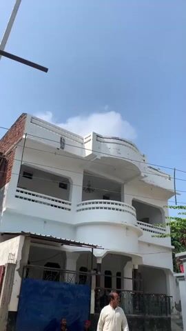 A man wearing a light-colored shalwar kameez walks to the right in front of a white, multi-story building with balconies. The building has a curved balcony on the second floor. In the background, there are trees and a bright blue sky with scattered clouds. A wire is visible running across the scene.