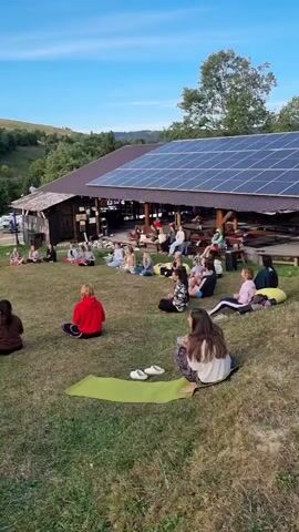 A group of people are sitting in a circle on a grassy lawn in front of a building with solar panels on the roof. Some people are sitting on yoga mats, while others are sitting on the grass. The building appears to be a rustic wooden structure with a covered patio area. In the background, there are rolling hills with trees and a clear blue sky. The overall scene suggests an outdoor gathering, possibly a yoga or meditation session, or a community event. The sun is shining, and the atmosphere appears peaceful and relaxed.