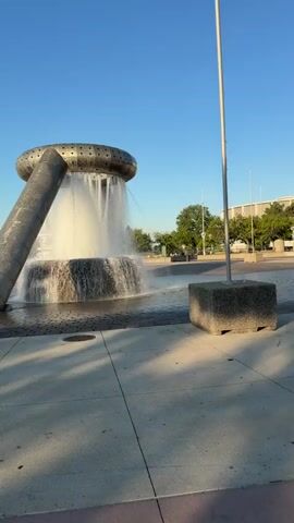 A fountain with water cascading from a metallic ring structure is in the foreground. A large cylindrical metal pipe supports the ring and arches over the water. The ground is paved with concrete and brick patterns. In the middle ground, a few people are walking along a promenade with trees and a city skyline in the distance. The sky is clear and blue.