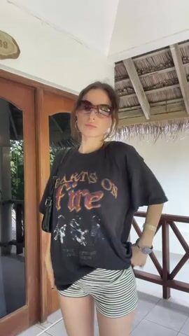 A young woman with brown hair pulled back stands in front of a wooden door with glass panels. She is wearing oversized sunglasses, a black t-shirt with a graphic print, and striped shorts. She holds her hands on her hips and slightly turns her body towards the camera. The background shows a thatched roof structure and a wooden railing. The video appears to be a short, candid clip.