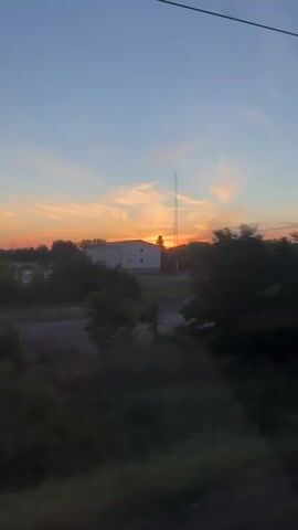 The video shows a sunset over a rural landscape. The sky is a vibrant gradient of blue to orange, with wispy clouds. Silhouetted trees and bushes are visible in the foreground, with a few buildings and a road in the mid-ground. The sun is partially obscured by trees, casting a warm glow. The camera appears to be moving, possibly from a vehicle, as evidenced by the slight blur and the occasional appearance of a wire or railing in the frame. The scene is peaceful and evokes a sense of the end of the day.