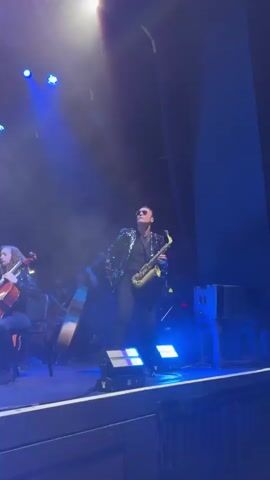 A man is playing a saxophone on a stage. He is wearing a black sequined jacket, a black shirt, and black pants. He is also wearing sunglasses. There are several other people on the stage, some of whom are playing musical instruments, including a cello and a double bass. The stage is lit with blue spotlights, and there is some smoke or haze in the air. The audience is not visible. The video is shot from a low angle, looking up at the performer.