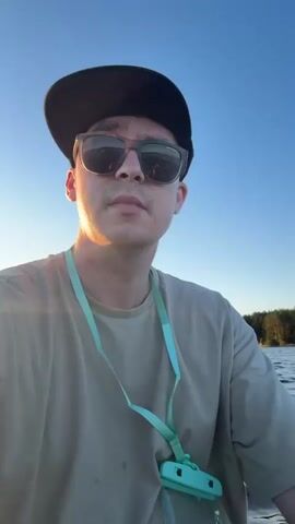 A man is taking a selfie while on a body of water. He is wearing a black baseball cap and sunglasses. He has a teal lanyard around his neck with a small case attached. The sun is low in the sky, casting a warm glow on the water and the man's face. In the background, there is a treeline and a clear blue sky. He appears to be on a paddleboard.