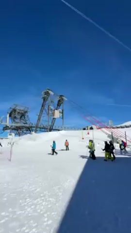 The video shows a snowy mountain scene with a ski lift infrastructure in the background. People are skiing and standing on the snow. There is a bright blue sky overhead with contrails from an airplane. The scene then shifts to show a wide view of snow-covered mountains. A person wearing a ski helmet and goggles stands near a safety net, looking out at the view and using a phone.