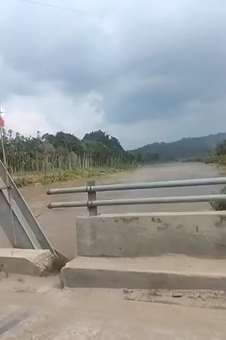 A video shows a muddy river flowing under a bridge. The river is lined with trees and lush vegetation on both sides. The sky is overcast with dark clouds, suggesting impending rain. The bridge has a concrete railing with metal bars, and the road leading up to it is also made of dirt and gravel. The camera pans slightly to the right, revealing more of the river and the surrounding landscape. The video appears to be filmed from a moving vehicle.
