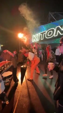 A group of people are on a stage at night. A large screen behind them displays the word "KITONE" in neon green letters. A person in the center of the stage wearing an orange jumpsuit and a white top is holding a device that is shooting flames. Another person in a dark shirt and sunglasses is standing to the left, holding their arms up. Other people are standing and watching or dancing. The stage is wet, reflecting the lights and flames. There are speakers on the stage.