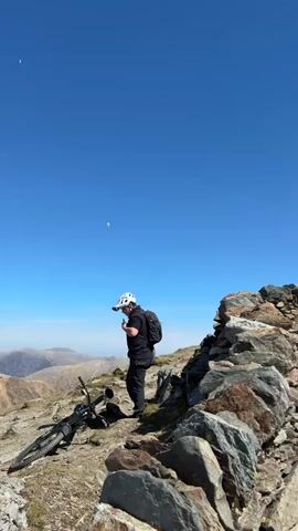 The video shows a rocky mountain summit with a clear blue sky.  A man with a bicycle and a white helmet stands on the left. A couple walks up stone steps. Many people are gathered at the top of the summit. A woman in a blue shirt with a backpack is visible in the foreground. A paraglider is visible in the distance.