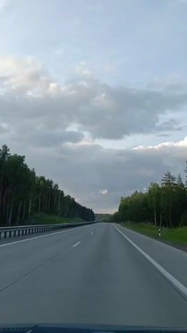 The video shows a forward-facing view from a car driving on a highway. The road is asphalt with white lane markings. On either side of the highway are dense forests with tall trees. The sky is mostly cloudy, with patches of blue visible. The lighting suggests it is either early morning or late afternoon. There are other vehicles visible in the distance on the highway. No people are visible in the frame. The car's dashboard is partially visible at the bottom of the frame.
