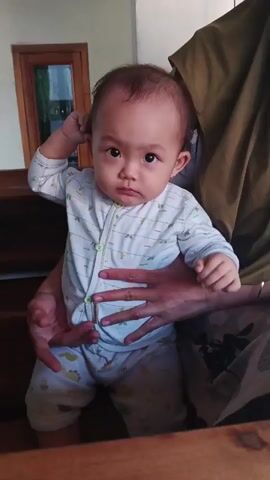 A baby is held by an adult and is looking at the camera. The baby is wearing a white onesie with green buttons and light blue pants with a dinosaur print. The adult is wearing a green headscarf. The background is out of focus but appears to be indoors with wooden furniture.