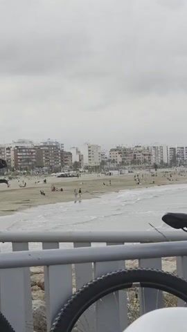A person filming from a vantage point is looking at a beach with a row of buildings behind it under a cloudy sky. The beach is populated with people walking along the shore, some lying on the sand, and others near beach bars. Waves are visible breaking on the shore, and a bicycle tire is in the foreground.
