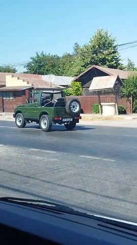 The video shows a green, open-top, off-road vehicle driving down a paved road. The vehicle has a spare tire mounted on the back. The interior of another vehicle is visible in the foreground, including a dashboard, steering wheel, and a person's hand on the steering wheel. The road is lined with houses and trees. The sky is clear and blue.