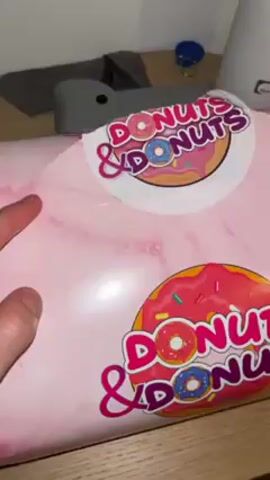 The video shows someone opening a pink box labeled "Donuts & Donuts" to reveal six donuts. Four are visible in the first shot, with a variety of toppings and icing, including a donut covered in colorful sprinkles and a "Carambar" candy on top of one of the donuts. The video then zooms out to reveal a total of six donuts.