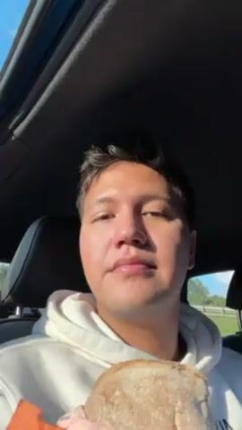 A man in a car is eating a sandwich wrapped in an orange napkin. The man is wearing a white sweater. He takes a bite of the sandwich and chews. We can see the inside of the sandwich and the road outside the car.